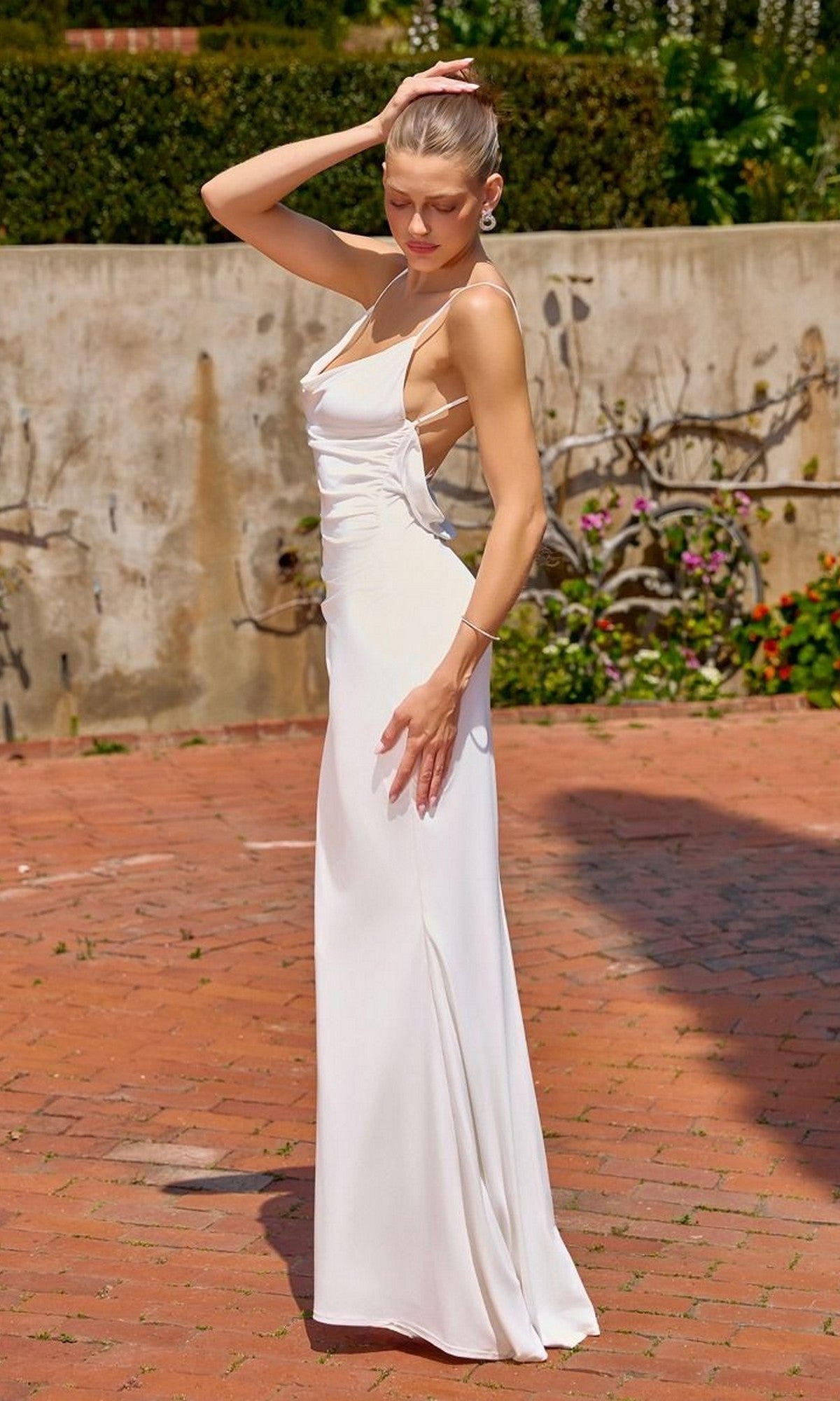 A woman wearing the Astee T2008W Long Formal Dress in off white stands outside on a red brick path, with one hand on her head and the other resting on her hip. Greenery and a stone wall are visible in the background.